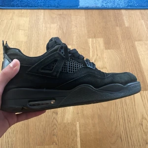 Svarta Nike Jordan 4 sneakers - Säljer ett par svarta Nike Jordan 4 sneakers med snygga meshdetaljer på sidorna och klassisk snörning. Skorna har en platt sula och är tillverkade i mocka med gummidetaljer. Perfekt för dig som gillar streetwear och vill ha en ikonisk look.