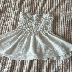 Vit bandeau kjol med volang - Supersöt vit bandeau kjol med smockad överdel och volangig nederkant. Kjolen är ärmlös och har en lätt strukturerad känsla i tyget, perfekt för varma dagar. 