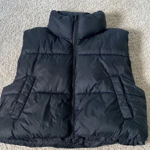 Svart dunväst från Pull&Bear - Säljer en svart, quiltad dunväst från Pull&Bear med hög krage och dragkedja framtill. Västen har två snedställda fickor och en stilren, puffig look som är perfekt för lager-på-lager. Passar dig som vill ha en enkel och snygg väst till höst och vinter.