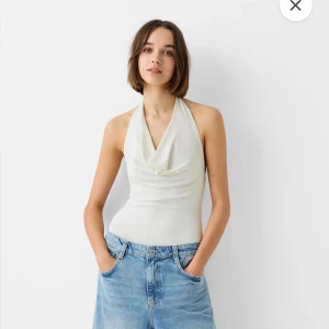 Vit omlott halterneck body  - Säljer en vit halterneck topp med omlott och draperad front. Toppen är ärmlös och har öppen rygg, vilket ger en snygg och fräsch look. Materialet är mjukt och stretchigt, troligtvis polyester eller syntet. Perfekt till jeans eller kjol för en cool sommarstil.