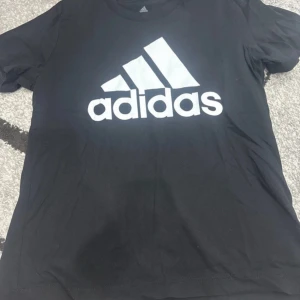 Svart Adidas t-shirt med logga - Svart t-shirt från Adidas med stor vit logga framtill. Klassisk rund hals och korta ärmar. Perfekt för dig som gillar sportig och enkel stil. Tillverkad i mjukt material som känns skönt mot huden.