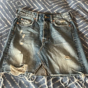 Blå jeanskjol med slitningar och fransar - Säljer en kort jeanskjol från H&M i ljusblå denim med coola slitningar och fransad nederkant. Kjolen har klassiska fickor fram och bak.💖