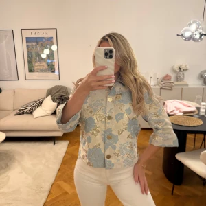 Populär blus från Vila - Säljer en superfin blus med stora blå och guldiga blommor på ljus botten. Blusen har har trekvartsärmar och snygga guldknappar framtill. Perfekt för dig som vill sticka ut med en elegant look.