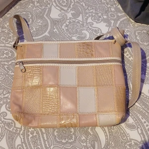 Beige och vit rutig axelväska i skinn - Snygg axelväska med patchwork-design i beige och vitt skinn. Rutigt mönster med olika texturer, vissa delar har reptilpräglad yta. Väskan har dragkedja upptill och ett orangefärgat innerfoder. Justerbar axelrem med dekorativa sömmar. Oanvänd.