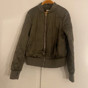 Grön bomberjacka från H&M - Snygg grön bomberjacka från H&M med quiltade ärmar och ribbade muddar. Jackan har dragkedja framtill och två fickor på sidorna. Perfekt för dig som gillar en stilren och avslappnad look.