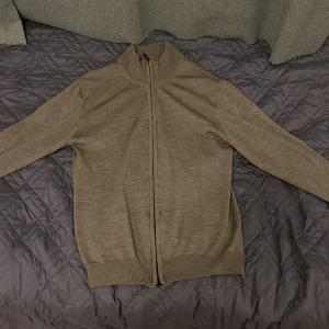 Beige kofta i ull från Sand Copenhagen - Snygg beige kofta från Sand Copenhagen i mjuk ull. Modellen har hel dragkedja framtill, hög krage och ribbade muddar vid ärmslut och nederkant. Perfekt för lager-på-lager och passar till det mesta. Enkel och stilren design.