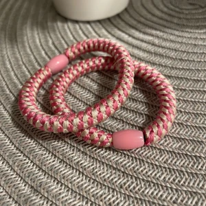 Randiga rosa och beige armband - Två chunky armband med randigt mönster i rosa och beige. De är gjorda av flätad syntet och har en rosa pärldetalj som avslut. Perfekt för dig som gillar färgglada accessoarer med lite retrovibe.