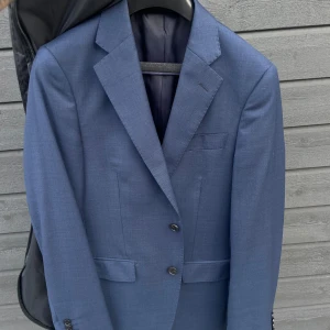 Blå kostym från These Glory Days - Snygg blå kostym från These Glory Days med klassisk kavaj och matchande byxor. Kavajen har två knappar, smala slag och flera fickor. Kostymen är helfärgad och har en stilren, modern passform. Perfekt för dig som vill ha en stilren look. Storlek 44 byxa och kavaj