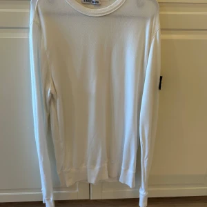 Vit långärmad tröja från Stone Island - Vit långärmad tröja från Stone Island med klassisk rund hals och ikonisk svart patch med logga på vänster ärm. Tröjan är tillverkad i mjuk bomull och har ribbade muddar vid ärmslut och nederkant. Perfekt för dig som gillar clean och stilren streetwear.