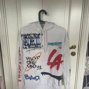 Vit hoodie med graffiti från Black Squad - Säljer en vit hoodie från Black Squad i relaxed fit, täckt av färgglada graffiti-prints och texter i blått, rött, svart och orange. Hoodien har huva med dragsko och långa ärmar. Perfekt för dig som gillar streetstyle och unika plagg.