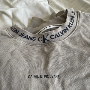 Beige sweatshirt från Calvin Klein Jeans - Säljer en beige sweatshirt från Calvin Klein Jeans med logga tryckt på bröstet och coolt CK-mönster runt kragen. Tröjan har en klassisk rund hals och är gjord i mjuk bomull, perfekt för chill dagar. Enkel och stilren design som funkar till allt.