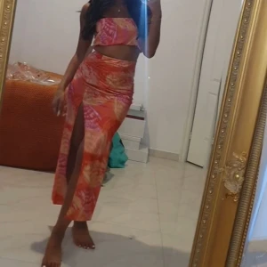 Färgglad bandeau tubtopp i orange och rosa och en kjol - Säljer en bandeau tubtopp med färgstarkt mönster i orange, rosa och röda toner. Toppen är ärmlös och axelbandslös, perfekt för varma dagar. Sitter tight och har en cool, somrig vibe. Matcha med kjol eller jeans för en trendig look.