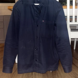 Mörkblå skjorta Tommy Hilfiger M - Snygg mörkblå skjorta från Tommy Hilfiger i storlek M. Klassisk design med knappar hela vägen, bröstficka med liten logga och krage. Tillverkad i mjuk bomull med lite stretch för extra komfort. Perfekt för dig som gillar stilrena och enkla plagg. Ny pris runt 2100