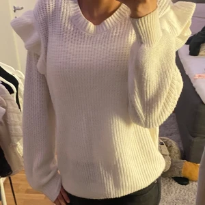 Vit stickad tröja - Superfin vit stickad tröja med breda volanger på axlarna och långärmad modell. Tröjan har en ribbad struktur och rund halsringning, perfekt för dig som vill ha något extra till din outfit.