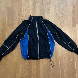 Svart och blå vindjacka vintage Dobsom - Sportig vindjacka från  i svart och blått med vita och gula detaljer längs ärmar och fram. Jackan har hög krage, dragkedja framtill, snörning i midjan och två fickor med dragkedja. Avtagbara armar