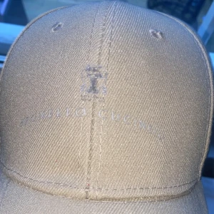 Beige keps från Brunello Cucinelli - Snygg beige keps från Brunello Cucinelli med diskret logga framtill. Klassisk form med böjd skärm och ventilerande hål upptill. Tillverkad i ett slitstarkt bomullsmaterial som ger en clean och stilren look. Perfekt för dig som gillar exklusiva accessoarer.