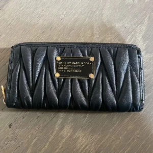 Svart quiltad plånbok Marc Jacobs - Snygg svart plånbok från Marc by Marc Jacobs med quiltad design och guldfärgade detaljer. Plånboken har dragkedja runtom, flera kortfack och ett myntfack med dragkedja. Märkesplakett i metall framtill ger en lyxig känsla. Se bild på slitningarna❤️