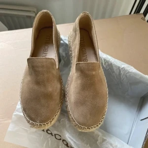 Beige mocka loafers från Macarena - Snygga loafers från Macarena i beige mocka med rund tå och platt sula. Skorna har en klassisk espadrille-look med grov söm runt sulan och är perfekta för dig som gillar enkel och stilren design. Passar till både jeans och shorts under varmare dagar.