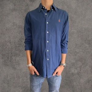 Mörkblå skjorta från Ralph Lauren S - Klassisk mörkblå skjorta från Ralph Lauren i bomull med custom fit. Skjortan har långa ärmar, knappar framtill och den ikoniska broderade loggan i orange på bröstet. Perfekt för en clean och stilren look.