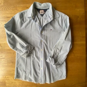 Blårandig skjorta från Tommy Hilfiger  - Snygg skjorta från Hilfiger Denim i blått och vitt med smala vertikala ränder. Klassisk krage, långa ärmar och knappar framtill. Liten broderad logga på bröstet. Tillverkad i mjuk bomull, perfekt för en chill och stilren look.