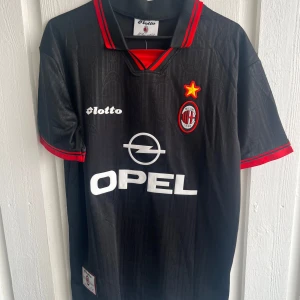 AC Milan Weah 14 svart fotbollströja - Säljer en klassisk AC Milan fotbollströja från Lotto med Weah och nummer 14 på ryggen. Tröjan är svart med röda detaljer på krage och ärmslut, samt vita tryck med Opel-logga och klubbmärke på bröstet. Materialet är lätt och andas, perfekt för match eller träning.
