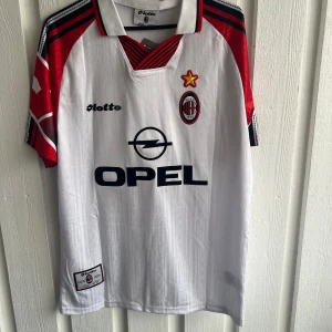 AC Milan Maldini fotbollströja Lotto - Säljer en klassisk AC Milan fotbollströja från Lotto med Maldini och nummer 3 på ryggen. Tröjan är vit med röda och svarta detaljer på ärmarna, har korta ärmar och krage med röd-svarta ränder. Materialet är lätt polyester, perfekt för match eller samling.