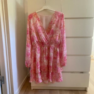 Blommig rosa kortklänning med volang - Säljer en superfin kortklänning med blommigt mönster i rosa och ljusa toner. Klänningen har omlott-v-ringad front, långa ärmar och volang ner till. Perfekt för sommarens alla tillfällen! Resår i midjan för skön passform. Han använda klänningen en gång på min kusins student och sen har jag glömt bort den, den e som ny och redo att användas!