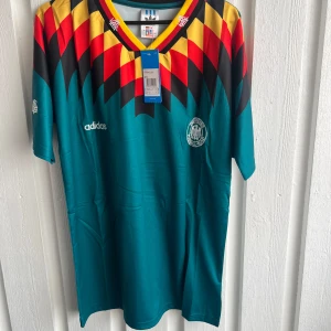 Tysklands retro fotbollströja Adidas - Säljer en ikonisk Tysklands retro fotbollströja från Adidas med MATTHÄUS och nummer 10 på ryggen. Tröjan är turkos med ett fett geometriskt mönster i rött, gult och svart över axlarna. Kortärmad och tillverkad i lätt polyester, perfekt för dig som gillar klassiska landslagströjor.