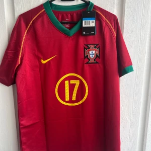 Portugal C. Ronaldo VM 2006 tröja - Säljer en officiell Portugal fotbollströja från VM 2006 med C. Ronaldo och nummer 17 på ryggen. Tröjan är röd med gula detaljer, gröna muddar och har landslagsemblem på bröstet. Tillverkad i lätt polyester med korta ärmar och snygg V-ringning.