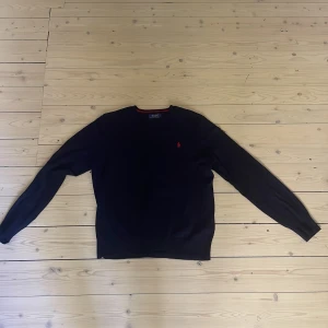 Mörkblå stickad tröja Polo Ralph Lauren - Snygg mörkblå stickad tröja från Polo Ralph Lauren med klassisk röd broderad logga på bröstet. Rund hals med röd detalj på insidan av kragen. Perfekt för dig som gillar stilrena och tidlösa plagg.