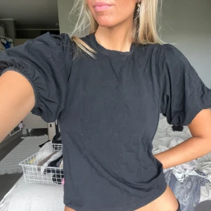 Svart t-shirt med puffärm - Svart t-shirt med coola puffärmar och rund hals. Modellen är lite loose och har en enkel, stilren look. Materialet känns som mjuk bomull och är perfekt för dig som gillar en chill vibe. Passar dig som vill ha något basic men ändå med en twist.