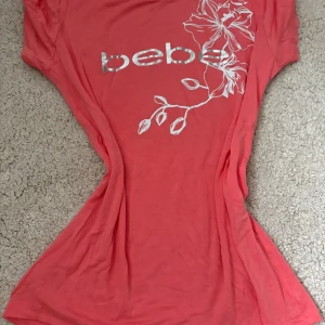 Korallrosa topp från Bebe med blommor - Snygg korallrosa topp från Bebe med silvrigt logotryck och vit blommig print på framsidan. Modellen är kortärmad och har rund hals. Materialet är mjukt och stretchigt med rayon och spandex, perfekt för en chill och trendig look. Oanvänd.