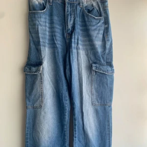 Blå cargopants från SHEIN, storlek M - Blå cargopants från SHEIN i medium med bred passform och stora fickor på sidorna. Jeansen har dragkedja och knapp framtill, klassisk femficksdesign och är gjorda i jeansmaterial. Perfekt för dig som gillar streetstyle och vill ha extra plats för prylar.