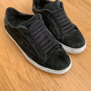 Svarta sneakers från Axel Arigato - Svarta sneakers från Axel Arigato med ovandel i mocka och detaljer i skinn. Skorna har svarta snören och vit platt sula med logotyp under. Stilren och minimalistisk design som passar till många olika looks.