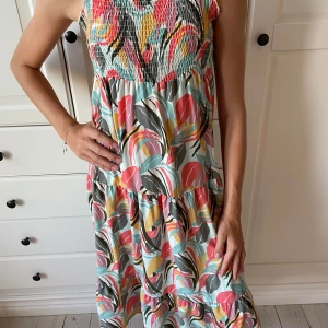 Färgglad långklänning från Name It - Säljer en superfin långklänning från Name It med smock upptill och axelbandslös design. Klänningen har ett färgstarkt blommigt mönster i rosa, gul, blå, grön och beige toner. Materialet är mjuk bomull, perfekt för varma dagar. Passformen är loose och klänningen har volangdetaljer. Går att spänna ut!