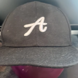 Svart keps med LA Airsport brodyr - Svart keps med vit broderad A på framsidan och en färgglad brodyr med texten 'Los Angeles Airsport 1982' på sidan. Justerbar snapback baktill. Materialet är bomull och kepsen har klassisk form med böjd skärm.