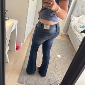 True Religion blå bootcut jeans - Säljer ett par snygga blå jeans från True Religion med klassiska röda sömmar och ikonisk bakficka. Modellen är bootcut med låg midja och har en cool faded effekt på låren. 