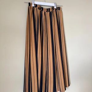 Randig långkjol i svart och beige - Säljer en flowy långkjol med svarta och beige vertikala ränder. Kjolen har resår i midjan för extra komfort och är gjord i 100% polyester. Perfekt för dig som gillar en chill och snygg stil med lite edge.