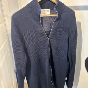 Mörkblå kofta från Selected Homme - Snygg mörkblå kofta från Selected Homme med dragkedja framtill och ribbad krage. Tröjan har ett coolt strukturerat mönster på bröstet och ärmarna, samt långärmad passform. Perfekt för dig som gillar stilrena och bekväma plagg.
