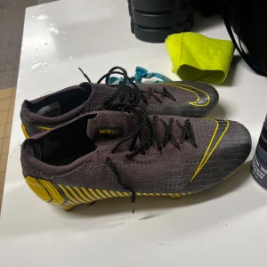 Nike Mercurial Vapor 360 fotbollsskor - Säljer ett par Nike Mercurial Vapor 360 fotbollsskor i mörkgrå och gul färg med snygga ränder och detaljer. Skorna har snörning och är tillverkade i syntetmaterial med en tight passform och platt sula med dobbar för gräsplan. Perfekt för dig som vill sticka ut på planen!