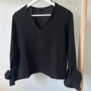 Svart blus med rosetter från Zara - Supersnygg svart blus från Zara Basic Collection med v-ringad hals och långa ärmar. Ärmsluten har stora rosetter. Blusen är lätt.