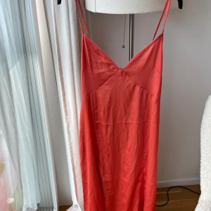 Orange långklänning från H&M med knyt - Säljer en orange långklänning från H&M i storlek XS. Klänningen har smala axelband, v-ringad fram och bak samt ett snyggt knyt i ryggen. Materialet är mjukt och har en lätt glansig finish som ger en lyxig känsla. Perfekt för dig som gillar stilrena och enkla plagg.