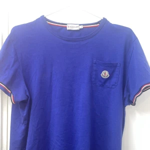 Blå Moncler t-shirt med ficka - Snygg blå t-shirt från Moncler med klassisk rund hals och korta ärmar. T-shirten har en bröstficka med Moncler-logga och coola röd-vita detaljer på ärmsluten och halsringningen. Tillverkad i mjuk bomull som känns skön mot huden.