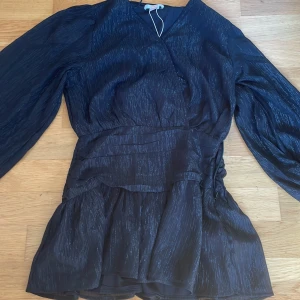 Svart glittrig kortklänning Gina Tricot - Supersnygg svart kortklänning från Gina Tricot med glittriga ränder i tyget. Klänningen har långärmade puffärmar, v-ringad hals och volang nedtill. Perfekt för dig som vill sticka ut med lite extra shine. Materialet känns lätt och har en lyxig glans.