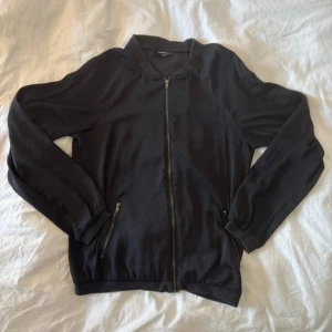 Svart jacka med dragkedja - Svart jacka från Kappahl med stilren design och hel dragkedja framtill. Jackan har två fickor med dragkedja och mjukt, lätt material som ger en skön passform. Perfekt att slänga på sig över en t-shirt eller hoodie för en chill look.