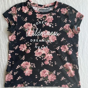Blommig svart t-shirt från KappAhl - Svart barn t-shirt från KappAhl med rosa blommigt mönster och vit text på magen. Rund hals, korta ärmar och gjord av bomull. Knappt använd och i mycket fint skick, hel och ren. Storlek 146/152