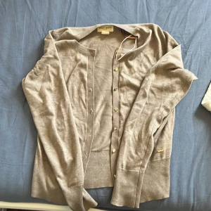 Beige kofta från Holly & White XS - Snygg beige kofta från Holly & White by Lindex i storlek XS. Den har rund hals, långa ärmar och guldfärgade knappar framtill. Materialet är mjukt och stickat, perfekt för lager-på-lager. Diskret metallmärke vid nederkanten ger en extra touch.