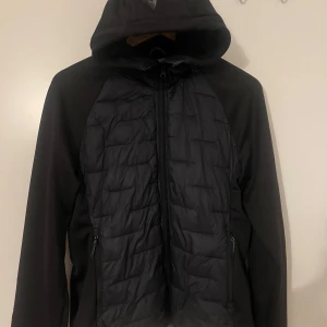 Svart mckinley jacka  - Svart pufferjacka från McKinley med huva och hel dragkedja. Jackan har quiltad front och rygg, släta ärmar och två sidofickor med dragkedja. Perfekt för dig som gillar sportig och clean stil. Materialet är syntet och polyester. Den är använd några gånger och är perfekt för regn o kallare väder. 