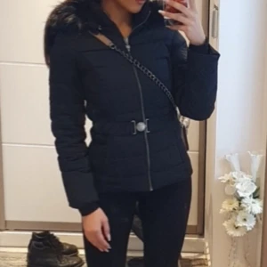 Svart pufferjacka med huva och bälte - Säljer en svart pufferjacka med snyggt bälte i midjan och dragkedja framtill. Jackan har en fluffig huva med pälsdetalj och långa ärmar. Perfekt för dig som vill ha en stilren och varm jacka med coola detaljer.