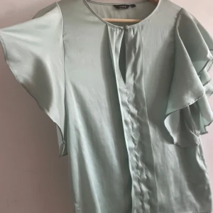 Ljusgrön blus med volang från Lindex - Supersöt ljusgrön blus från Lindex med volangärmar och rund halsringning. Blusen har en knappdetalj framtill och är tillverkad i ett glansigt, mjukt material som ger en lyxig känsla. Perfekt för dig som gillar flowy vibes och vill sticka ut med en fräsch färg.